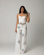 A woman takes a mirror selfie wearing a matching two-piece outfit consisting of a white corset-style crop top and white wide-leg pants with blue and green botanical patterns. She is standing in front of a brown hexagonal patterned wall.
