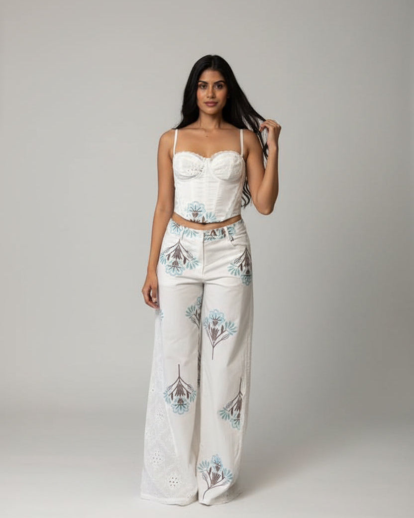 A woman takes a mirror selfie wearing a matching two-piece outfit consisting of a white corset-style crop top and white wide-leg pants with blue and green botanical patterns. She is standing in front of a brown hexagonal patterned wall.