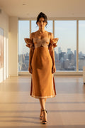 A woman modeling a rust-orange midi dress with a cream geometric spiral pattern on the bodice and sleeve cuffs, styled with black strappy high-heeled sandals against a plain white studio background.